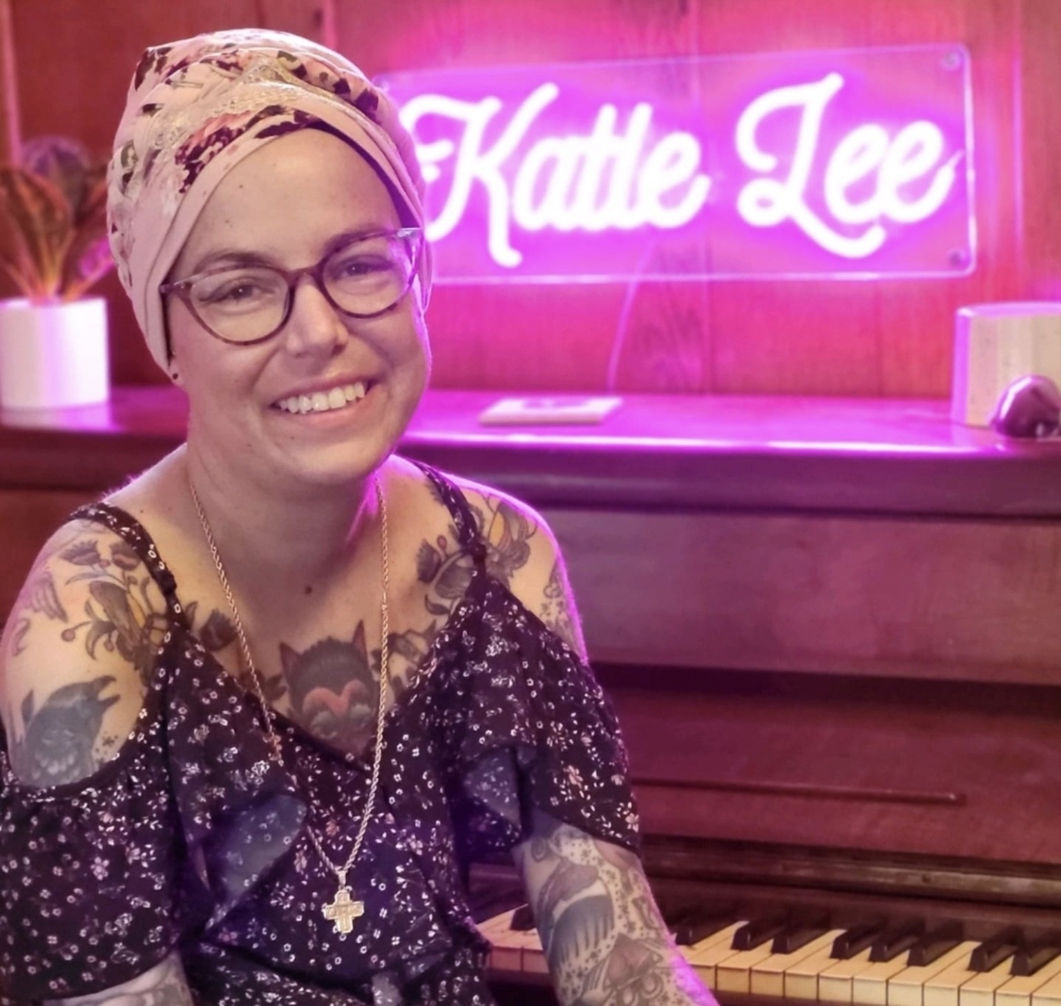 Katie Lee smiling at a piano, surrounded by her tattoos and a neon Katie Lee sign